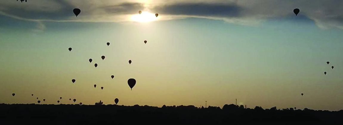 Silhouetted hot air balloons float across a dusky sky.