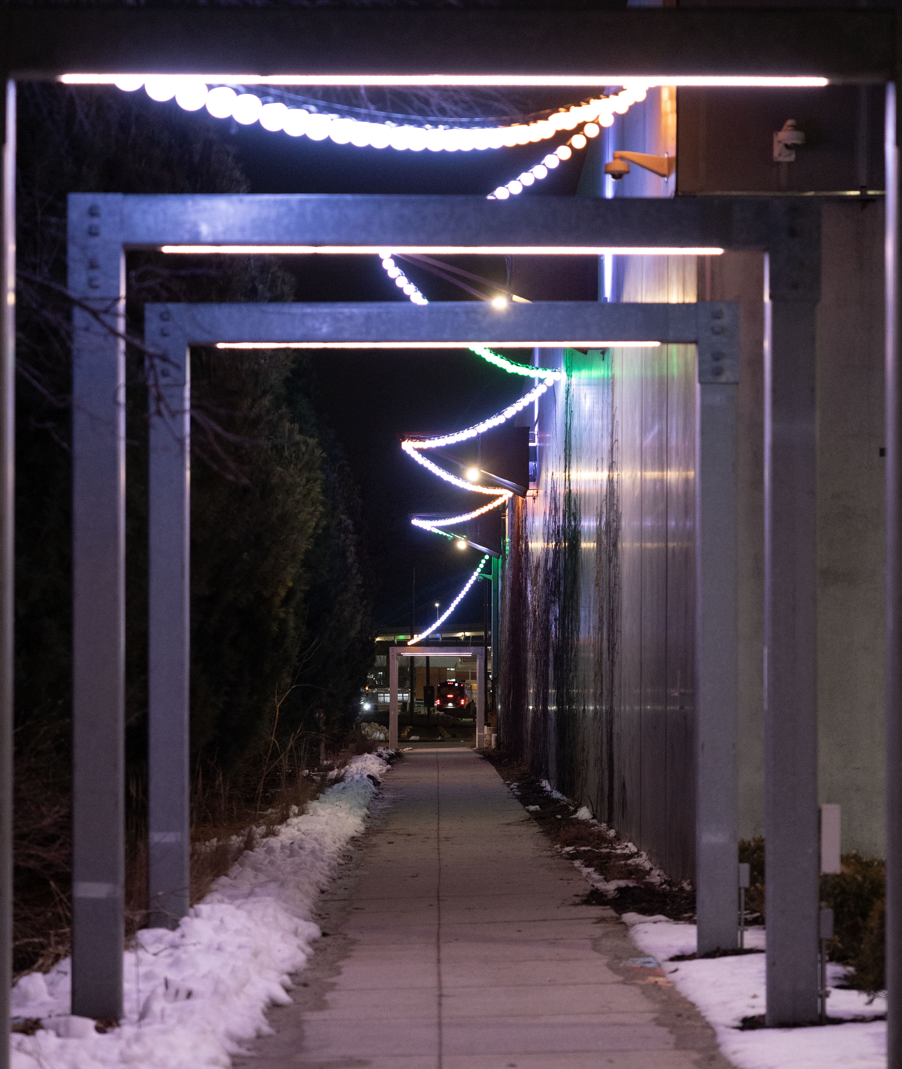 A pathway at night with three arches over the path and balled lights strung at the top. There are trees to the left and a building to the right. 