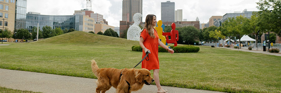 Woman walking her dog through the park with the Downtown Des Moines skyline in the background. 